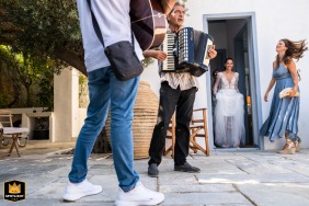 At The Anchor House in Symi, Greece, a WPJA wedding photographer captures a candid moment. A bridesmaid tidies the bride's hair as local Greek musicians perform in front of the house.